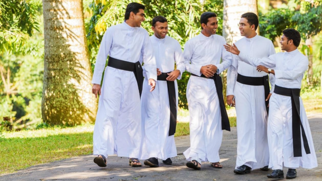 A group of young men in Seminarian cassocks walking along a sunny path, smiling as they talk to each other.
